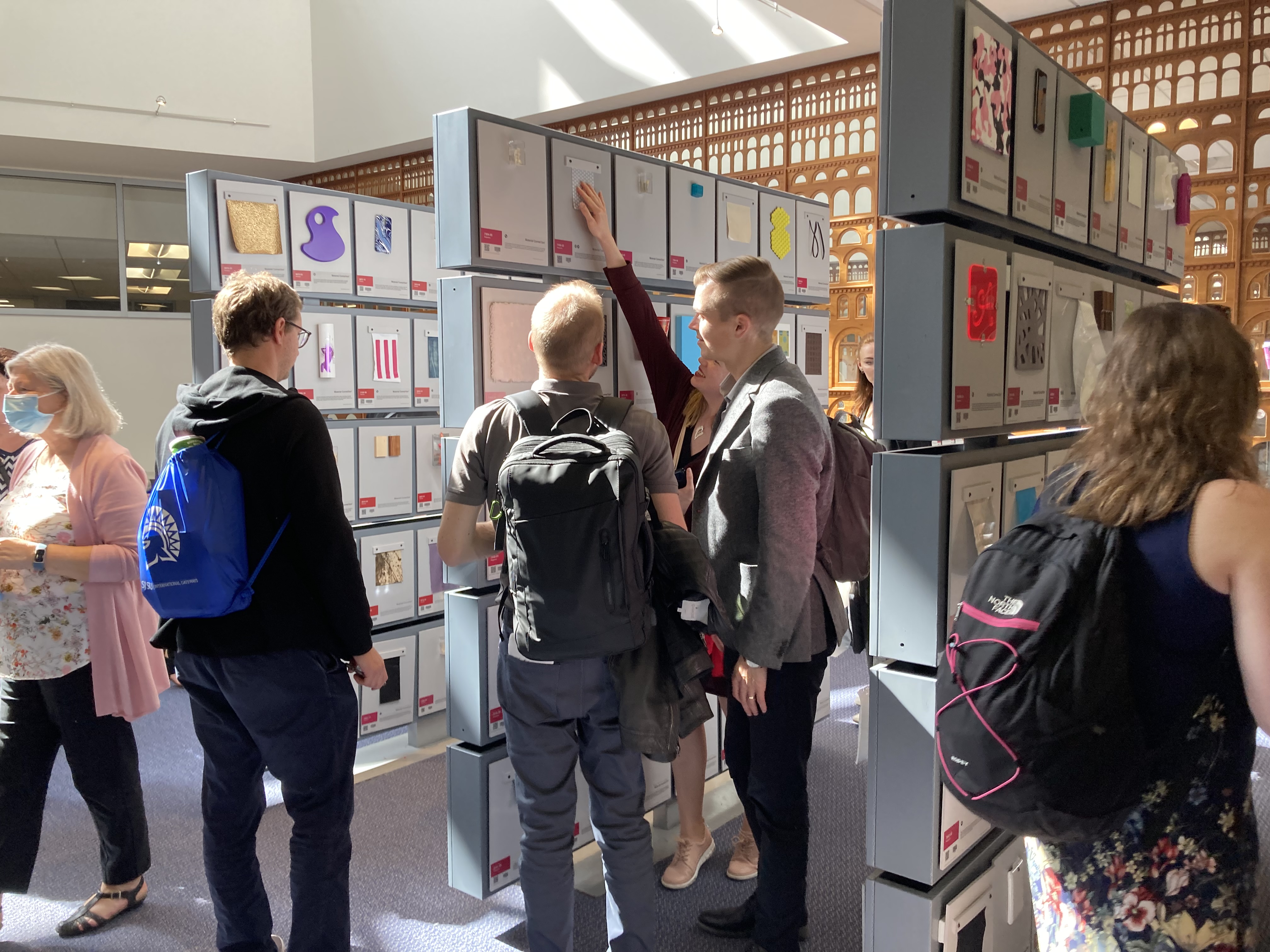 Three male students and one female stand in front of a panel of squares, each with a 4' scrap of plastic, fabric, or other material in SJSU's Materials Library.  A staff member with gray hair and a pink sweater walks away in the background, while a woman with a background walks to the right in the foreground.