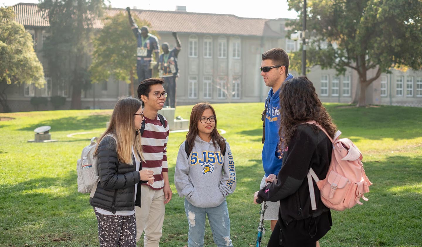 Five students talking on San José State University’s Tower Lawn with the Vicotry Salute monument in the distance.