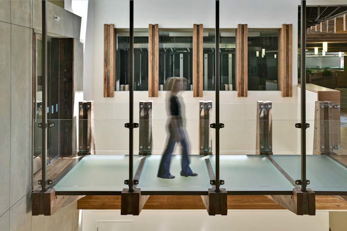 A person in motion walking on a glass bridge held up by metal rods and wooden beams.