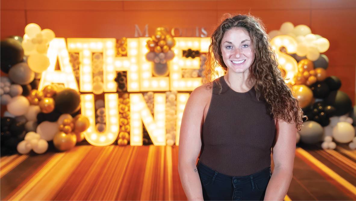 Alexa Solomon poses in front of a huge lit up sign that says Athletes United.