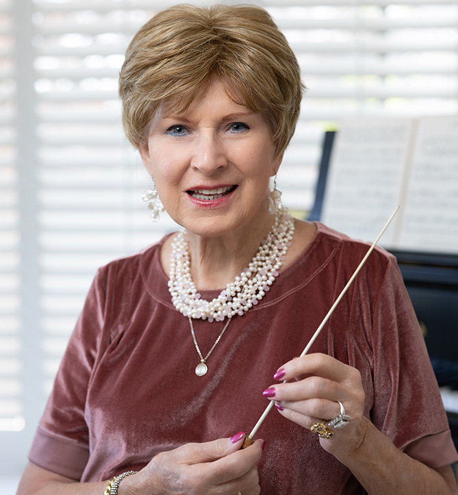 Woman in maroon top smiles in front of a piano, holding a conductor's wand. Charlene Archibeque, conductor, music, choir, professor emerita