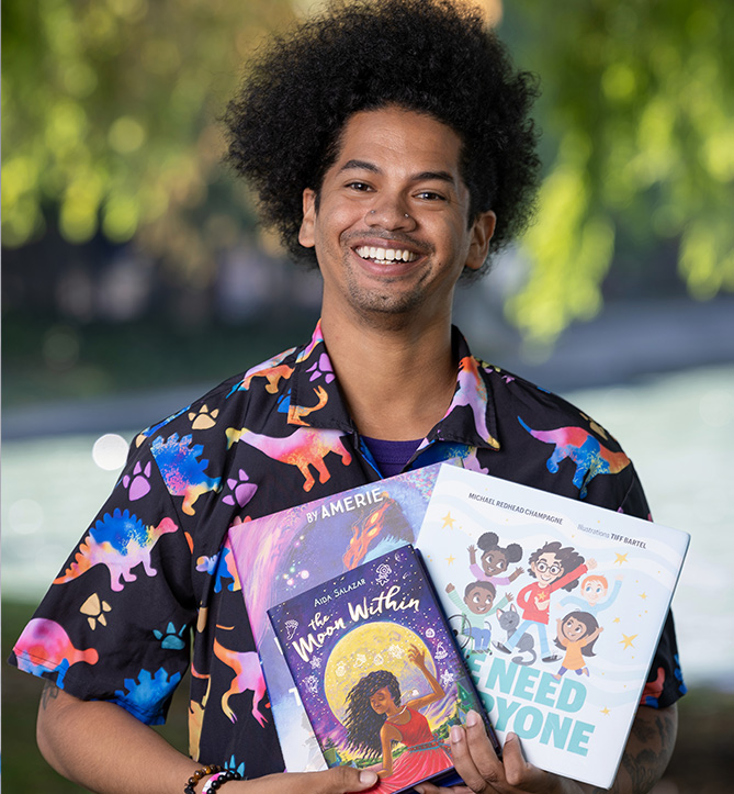 A smiley African American man in a t-shirt patterned with dinosaurs holding children's books faces the camera. Mychal Threets, literacy, graduate student, picture books