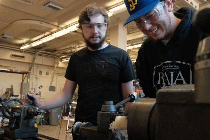 two students work over a lathe in the ME Machine shop. One is smiling at the work while the other observes the work intently.
