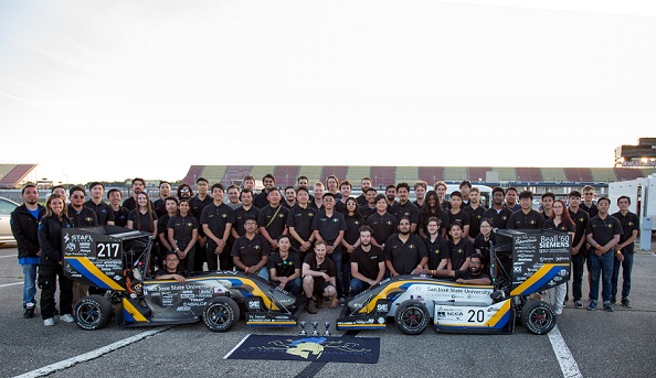 color photo of SJSU Spartan Racing team at the Michigan SAE competition. Both the Combustion and Electric Cars are in front of the group, with their drivers in the cockpit