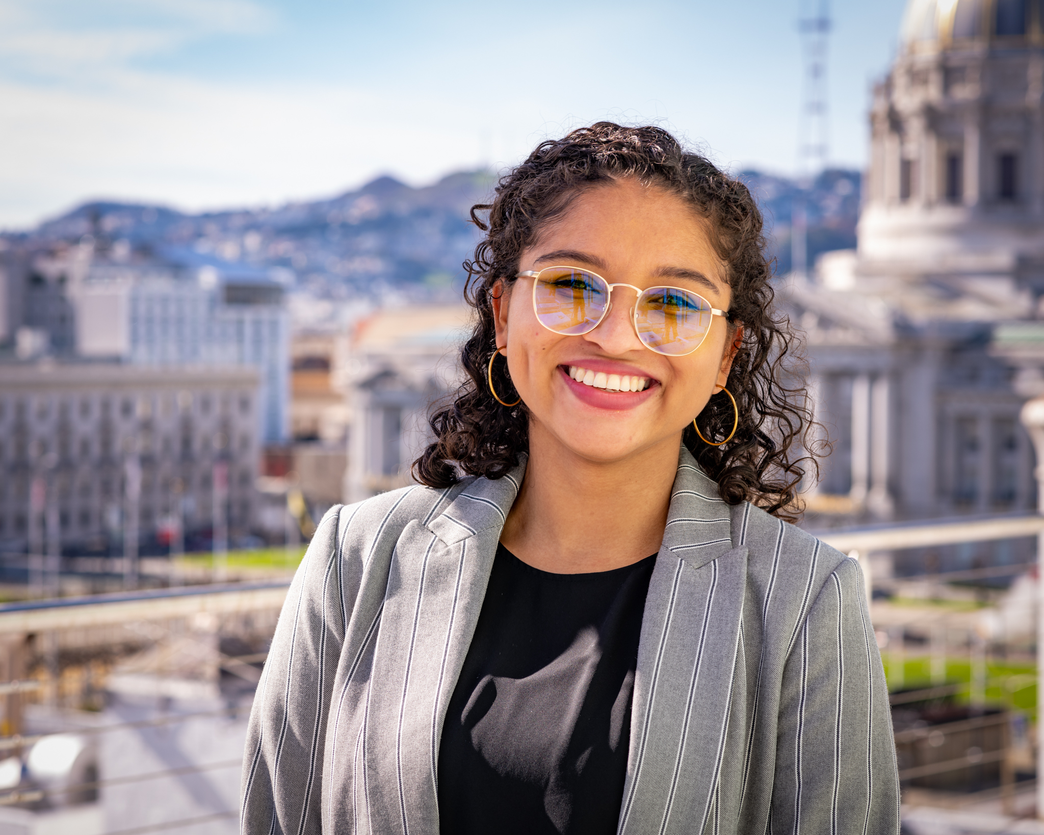 Hispanic woman with long, curly brown hair, wearing a gray suit jacket and black shirt, smiling in front of a state or federal capital building. 