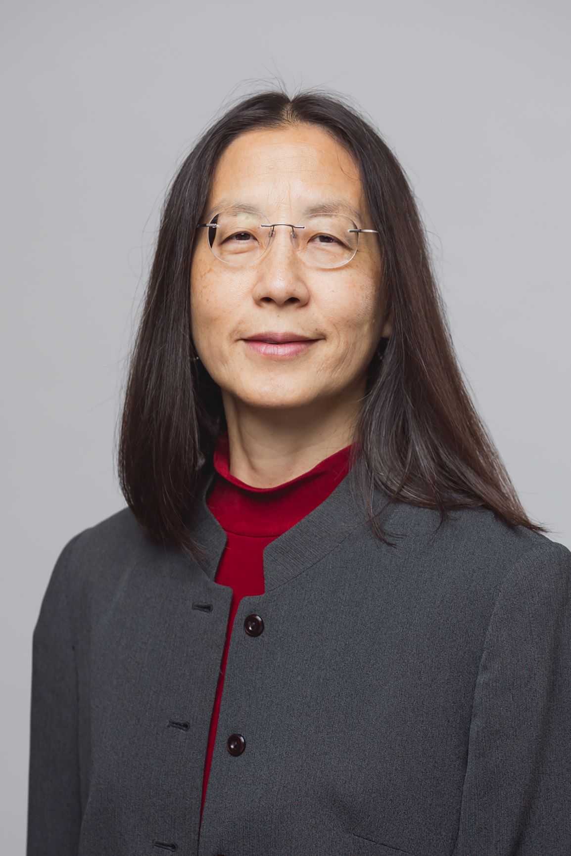 Asian woman with long, black hair, with rounded silver glasses, wearing a gray suit jacket and red shirt, smiling in front of a gray background.