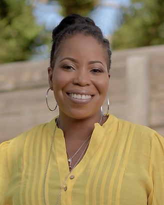 Black woman with tied-back, black hair, wearing a yellow button down, in front of a brown fence and green tree.