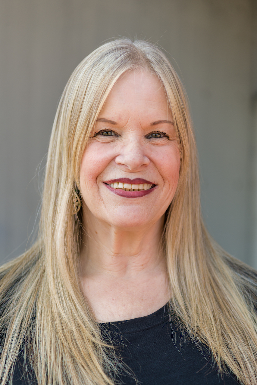 White woman with long, blond hair, with a black shirt and gold earrings smiling in front of gray background