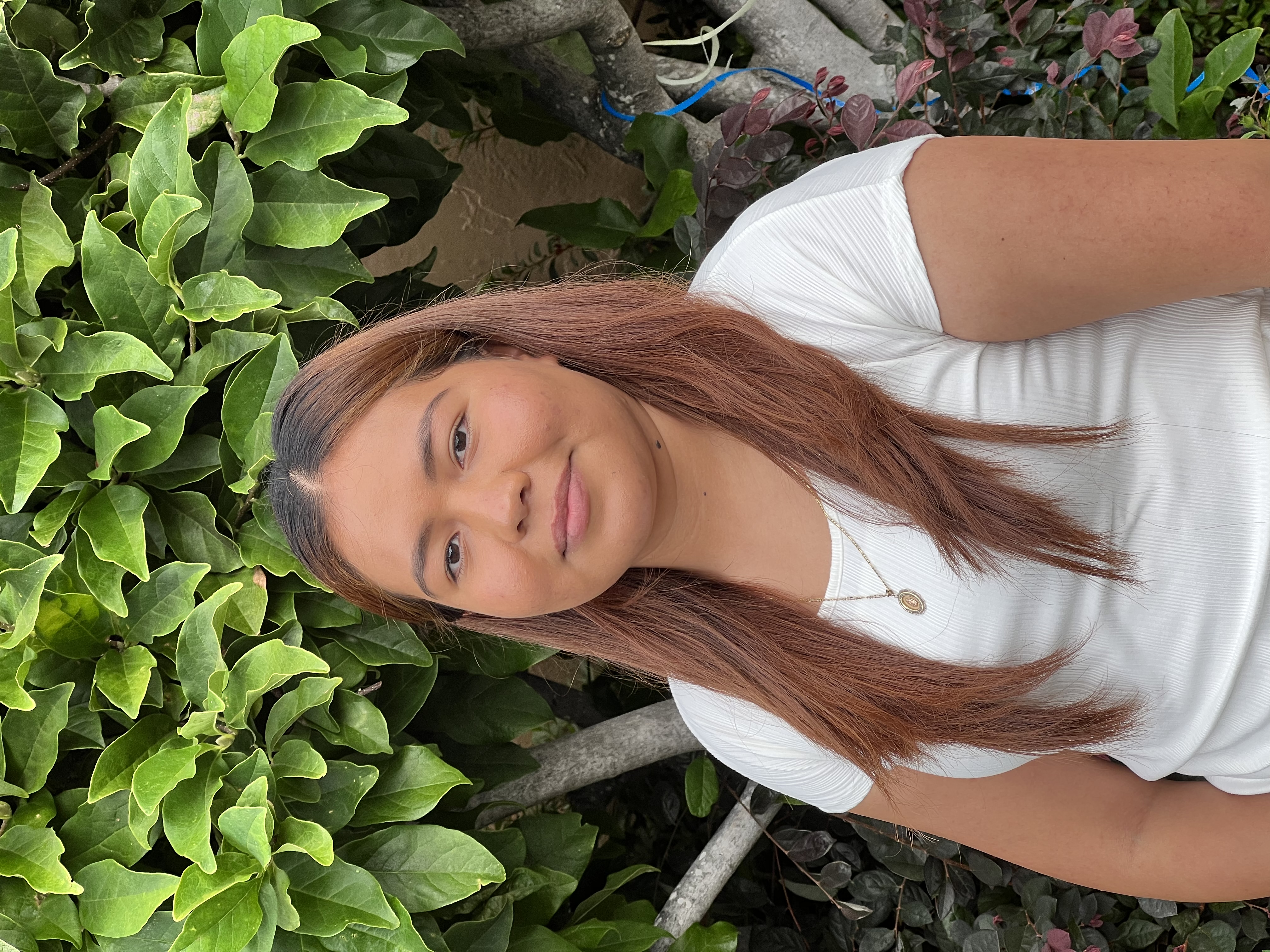 Hispanic woman in her twenties, with long, brown hair, wearing a white t-shirt and gold necklace smiling in front of green tree.