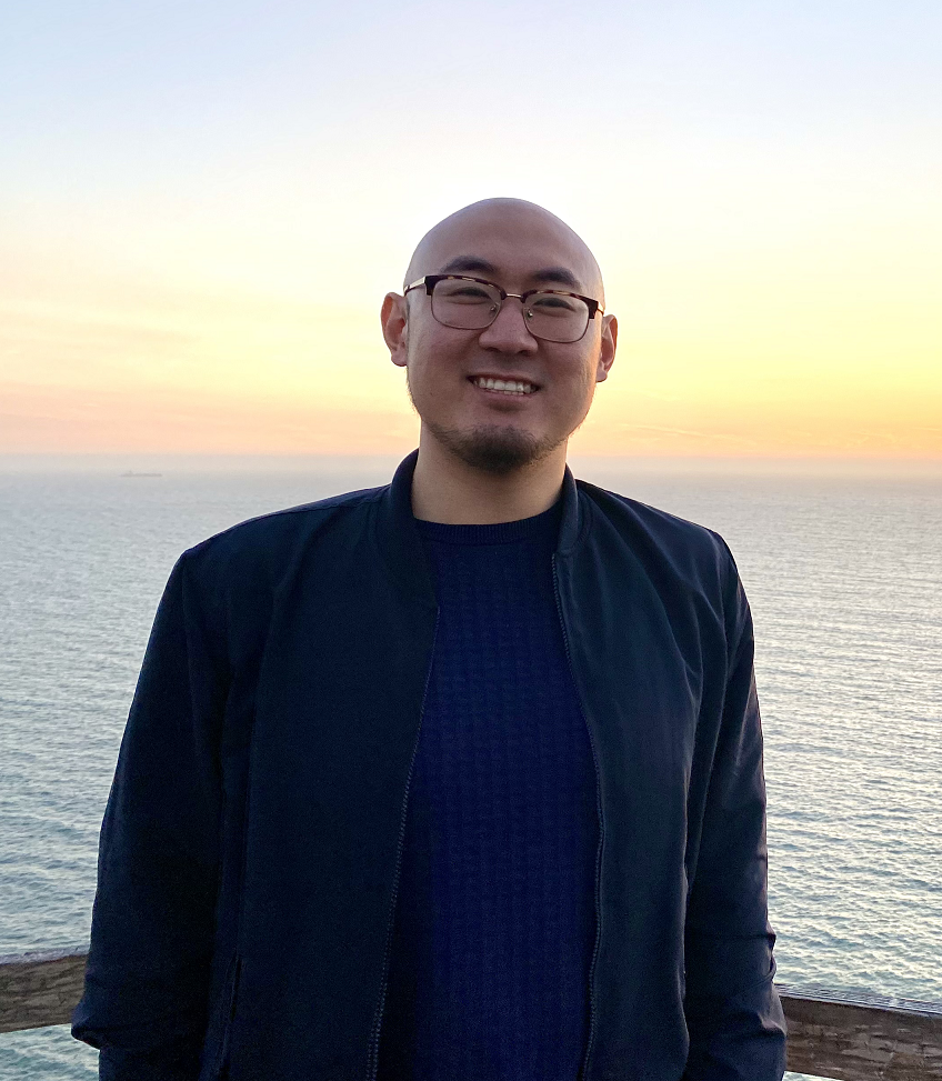 Asian male attorney, bald, wearing a black jacket and blue shirt, smiling in front of the ocean and a red-orange sunset.