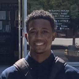 Black male in his twenties, with short, curly hair, wearing a black long-sleeve shirt standing in front of a storefront. 