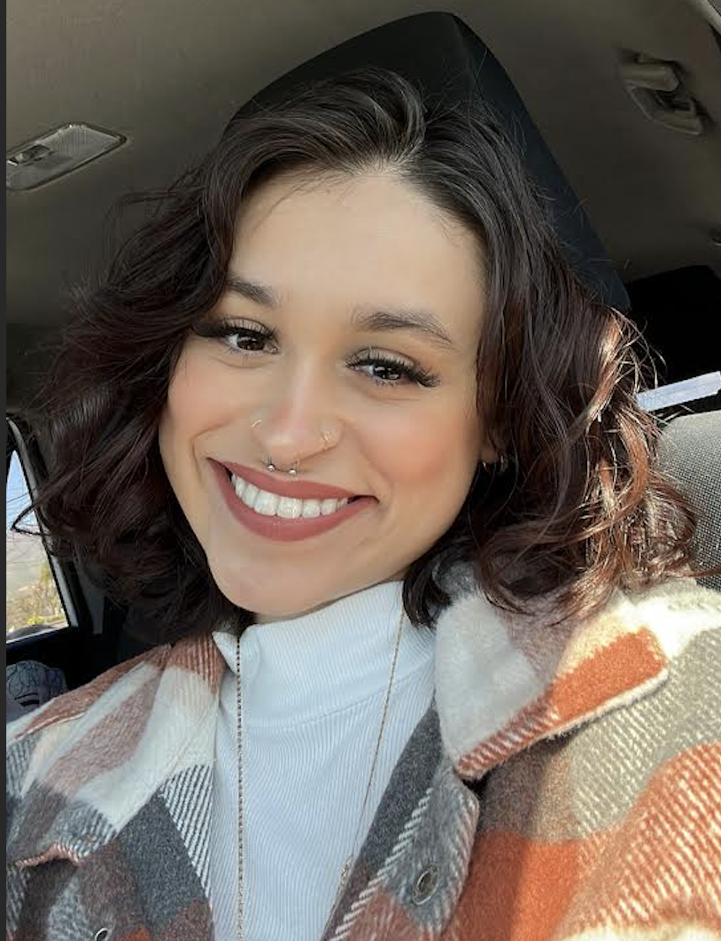 Hispanic woman in her twenties, with medium-length, short brown hair, wearing an orange and white checkered coat, smiling in the drivers seat of a car.