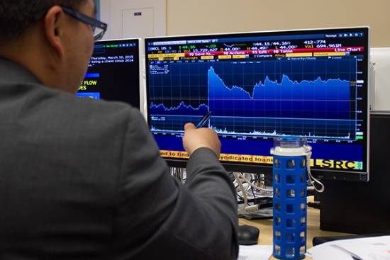 looking over the shoulder of a person in a suit, we see them pointing to a screen showing an economic chart