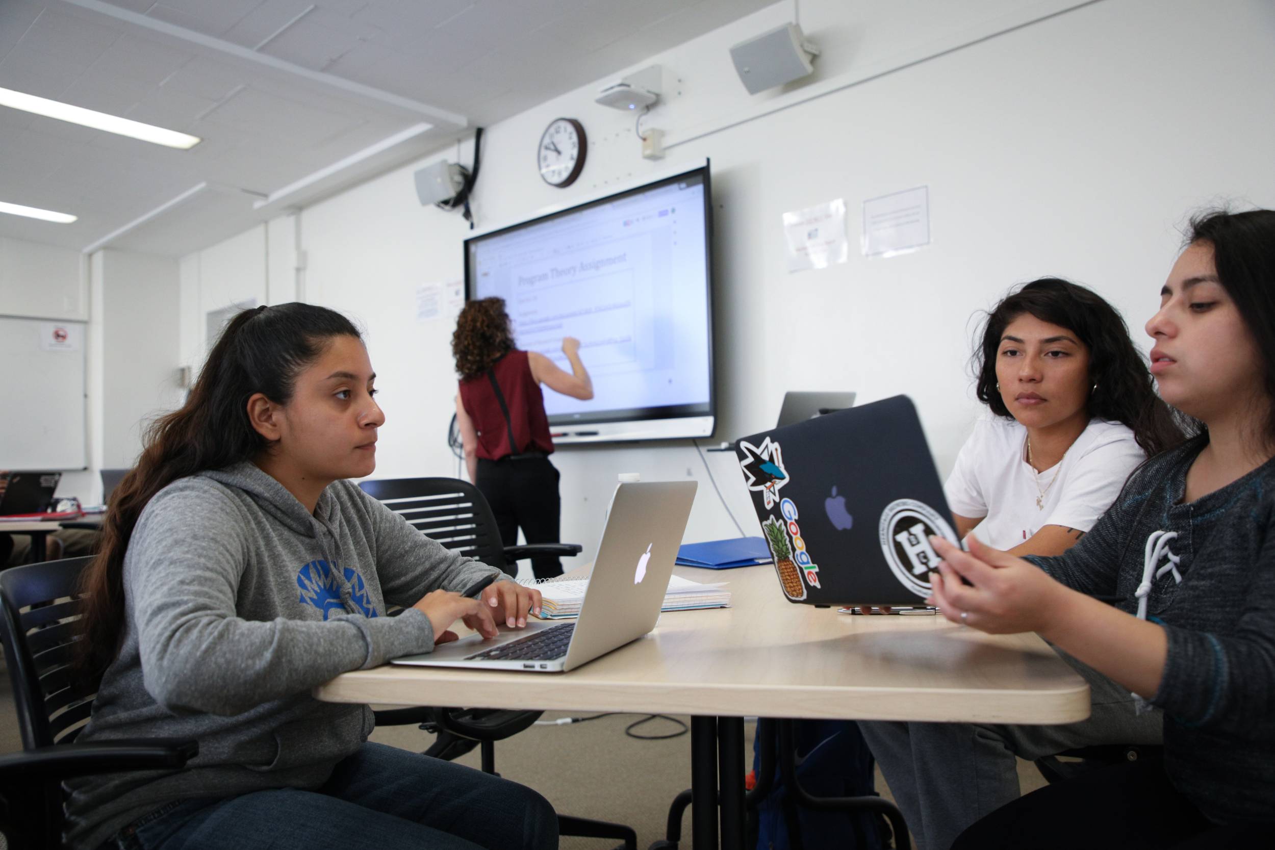 A group of students sit at a table working on laptops and discussing their work while a teacher teaches on a smart board in the background. 