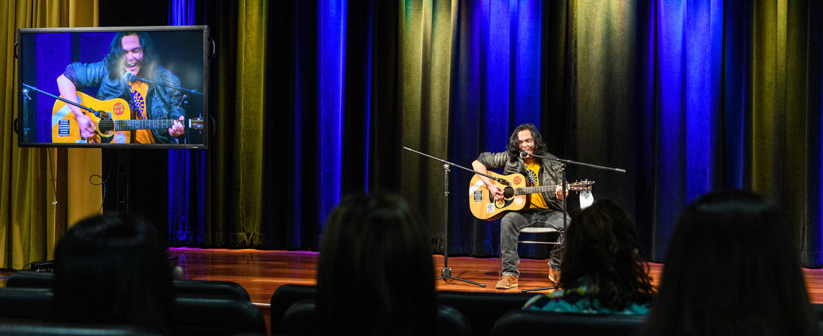 A student sings as he plays the guitar in a theatre lit up with blue and gold lights.